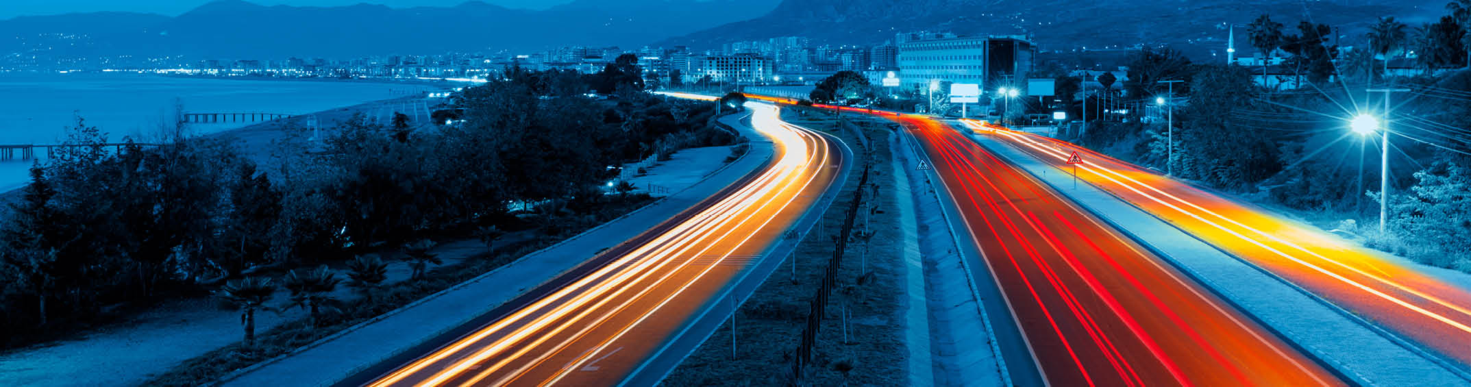 Lunga esposizione di un'autostrada di notte con scie luminose di veicoli che creano linee rosse e bianche mentre attraversano un paesaggio urbano vicino al mare. Descrizione generata da AI